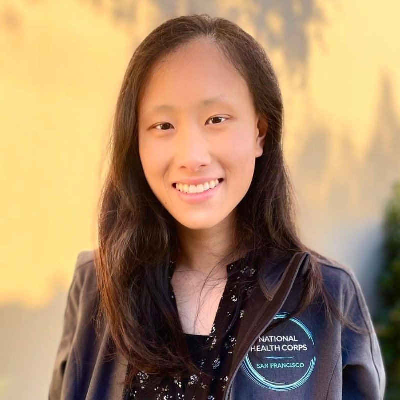Mindy is facing the camera smiling, wearing a charcoal grey National Health Corps jacket over a blouse. Behind her is a cream-colored, blank wall.