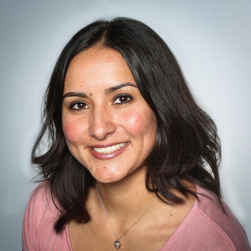 Lily Morales is seen here facing the camera and smiling. Her photo was taken against a grey white background. She is wearing a wide neck pink blouse.