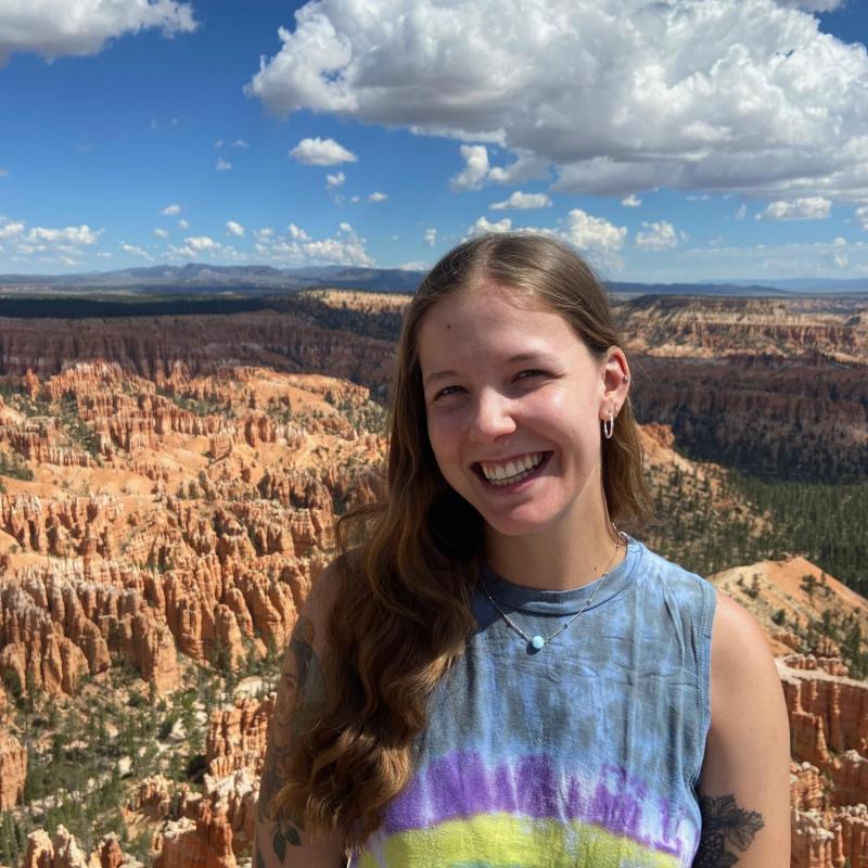 Grace Lemke is seen standing, facing the camera, smiling while wearing a tie-dyed tank top. 