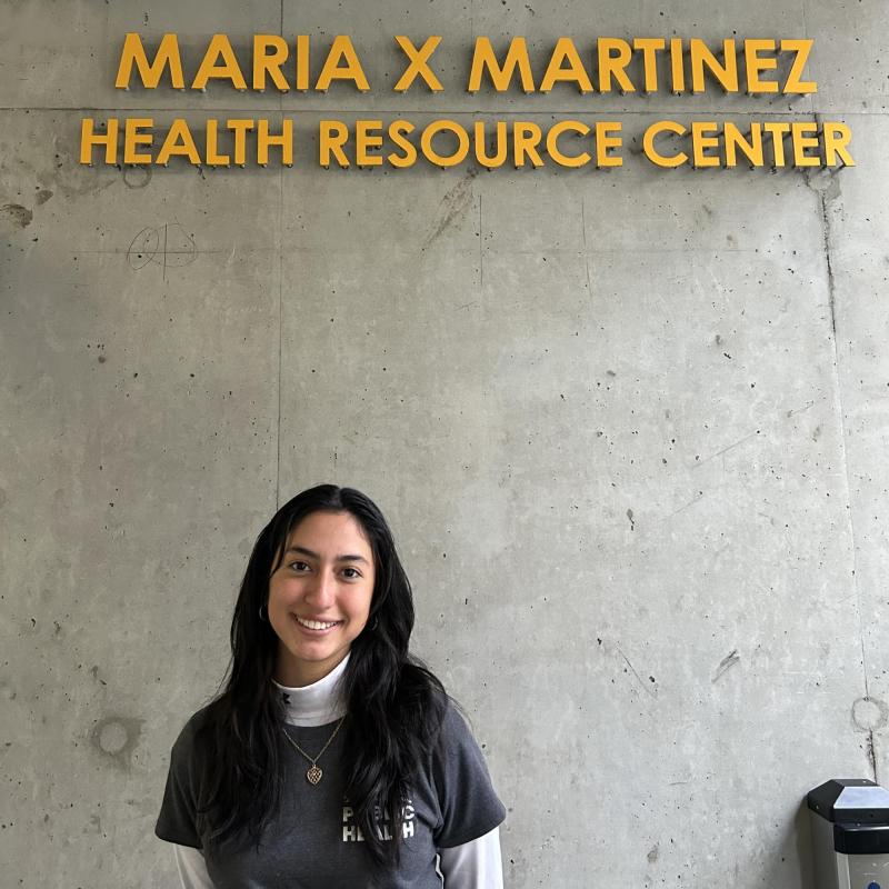 Laura is standing off center frame, facing the camera and smiling. Above her is the entrance sign for the Maria X Martinez Health Resource Center.